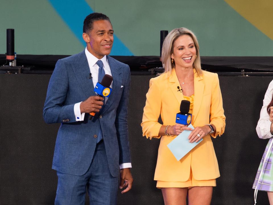 T.J. Holmes, in a blue suit, smiles while standing next to fellow "GMA" host Amy Robach, in a yellow blazer and skirt, on stage.