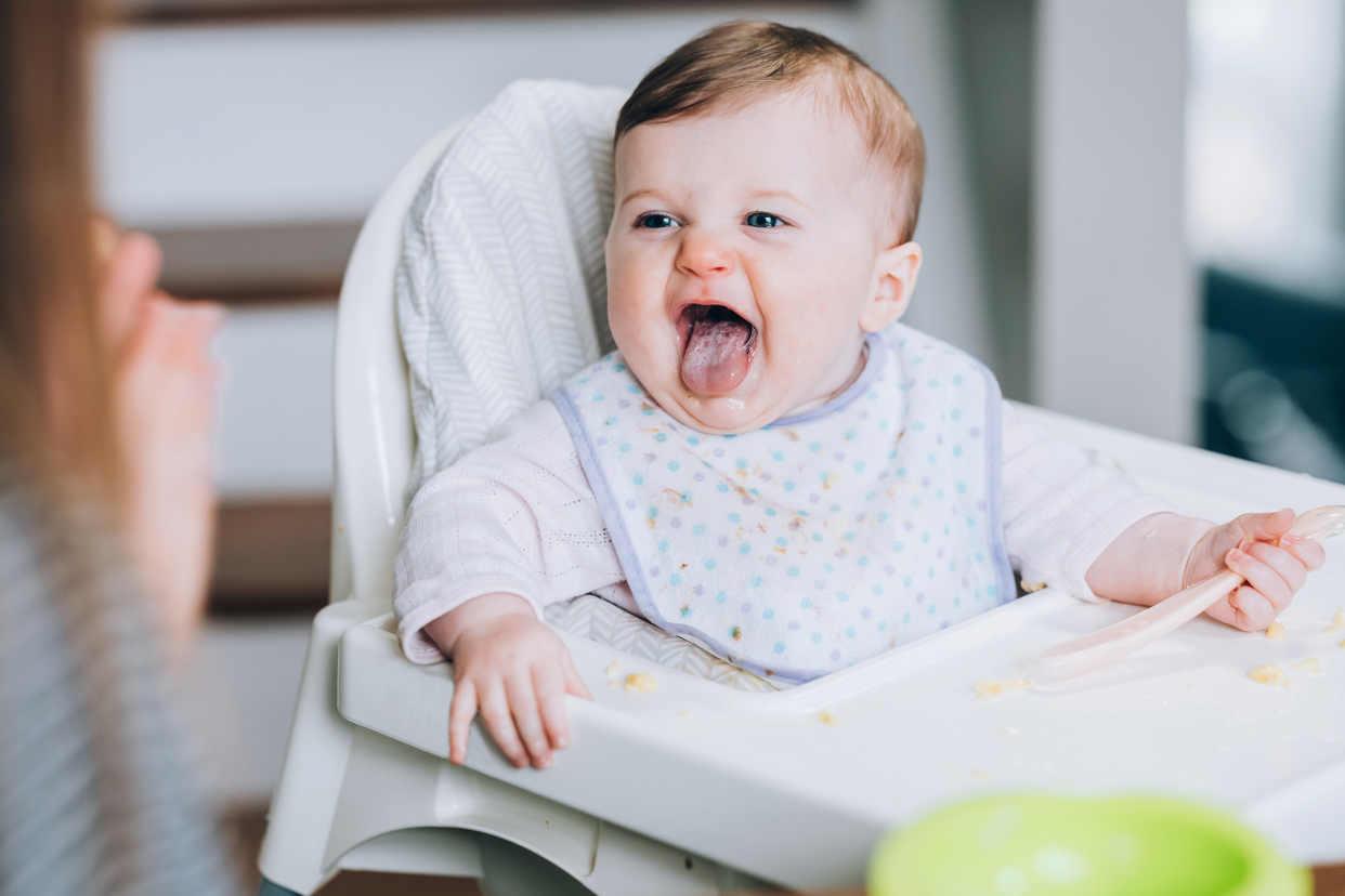 Baby girl making a happy face while being fed in a high chair, selective focus, looking at mother off the left side, modern kitchen blurred in the background