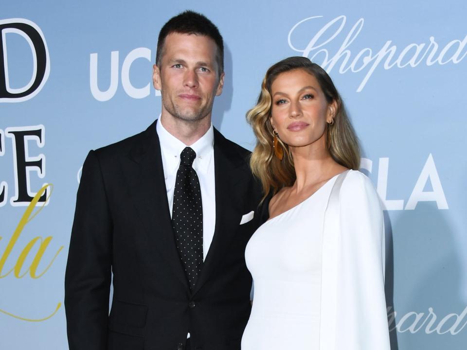 Tom Brady in black suit and Gisele Bündchen in fancy white dress in front of blue backdrop