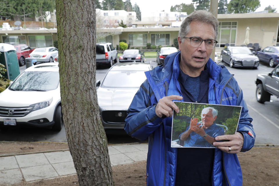 In this March 12, 2020, photo, Scott Sedlacek poses for a photo while holding a photo of his father, Chuck, outside Life Care Center in Kirkland, Wash., near Seattle. The facility has been at the center of the coronavirus outbreak in the state, and Sedlacek — who also has tested positive for the virus — said he and his siblings have barely spoken to their father, who in addition to testing positive for the coronavirus, has blindness, neuropathy, and has difficulty using a phone, saying he is more of an "inmate" than a patient. Residents of assisted living facilities and their loved ones are facing a grim situation as the coronavirus spreads across the country, placing elderly people especially at risk. (AP Photo/Ted S. Warren)