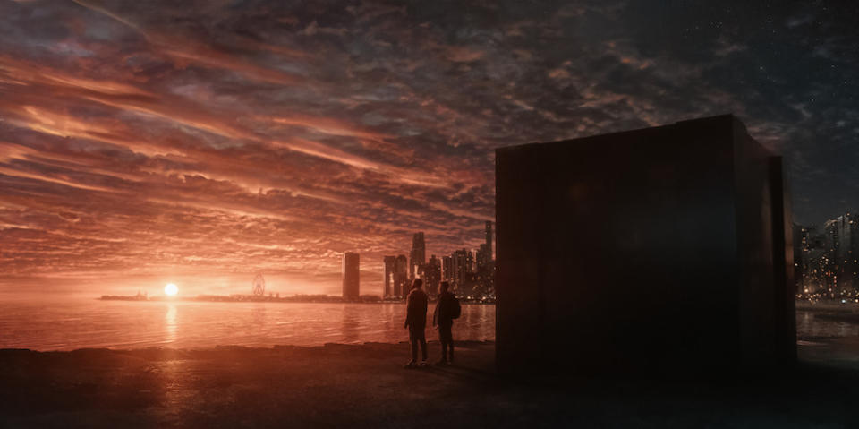 An image from the 'Dark Matter' TV series, showing two people standing next to a 12-foot tall black cube, sitting on a beach, overlooking Chicago at sunset