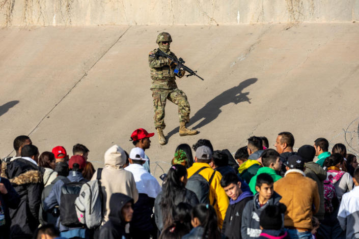 An armed Texas National Guard soldier blocks a crowd of migrants from entering an illegal border crossing area.
