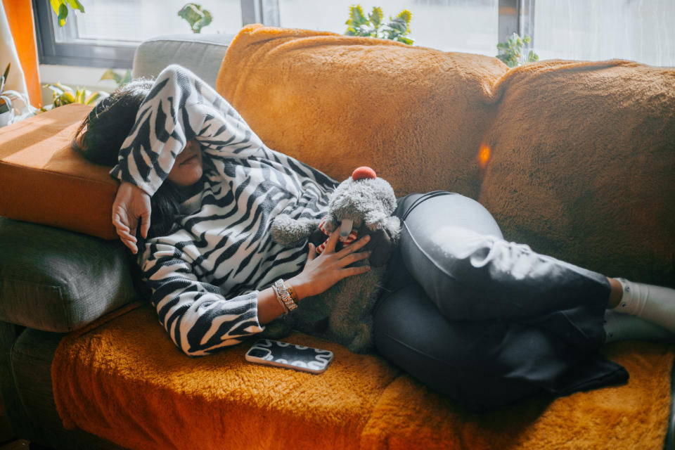 Person resting on a couch with a zebra-patterned sweater, arm over face, holding a teddy bear. A phone and an orange cushion are beside them