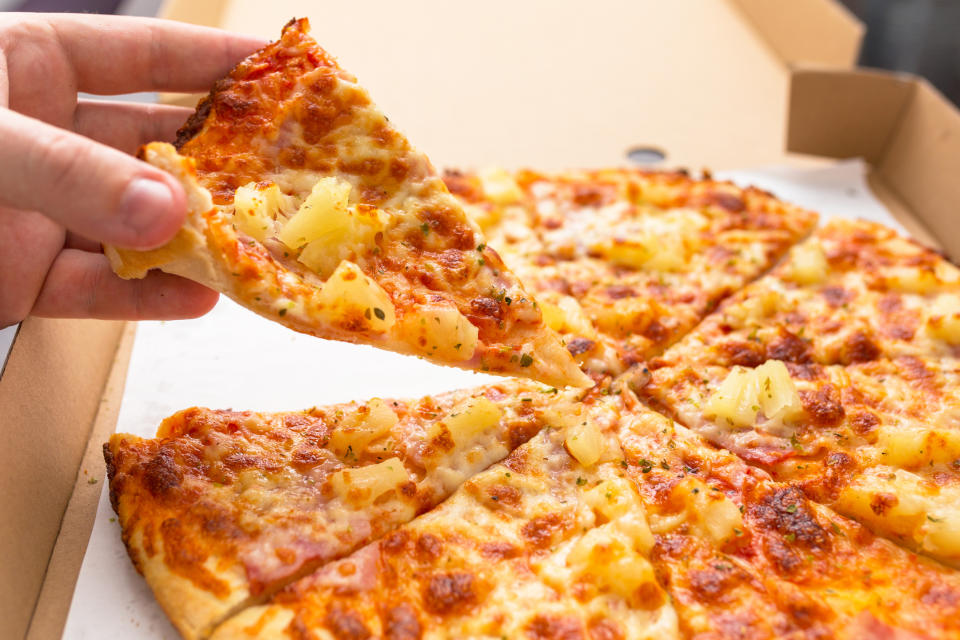 Hand holding a slice of pizza with pineapple and ham topping, taken from an opened pizza box