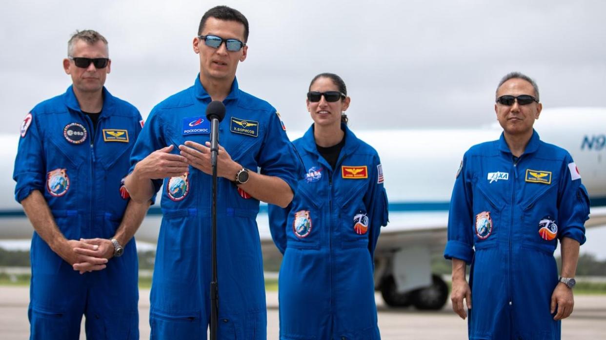  four astronauts in a row with flight suits on. one of them speaks at a microphone while three stand in the background. all are wearing sunglasses 