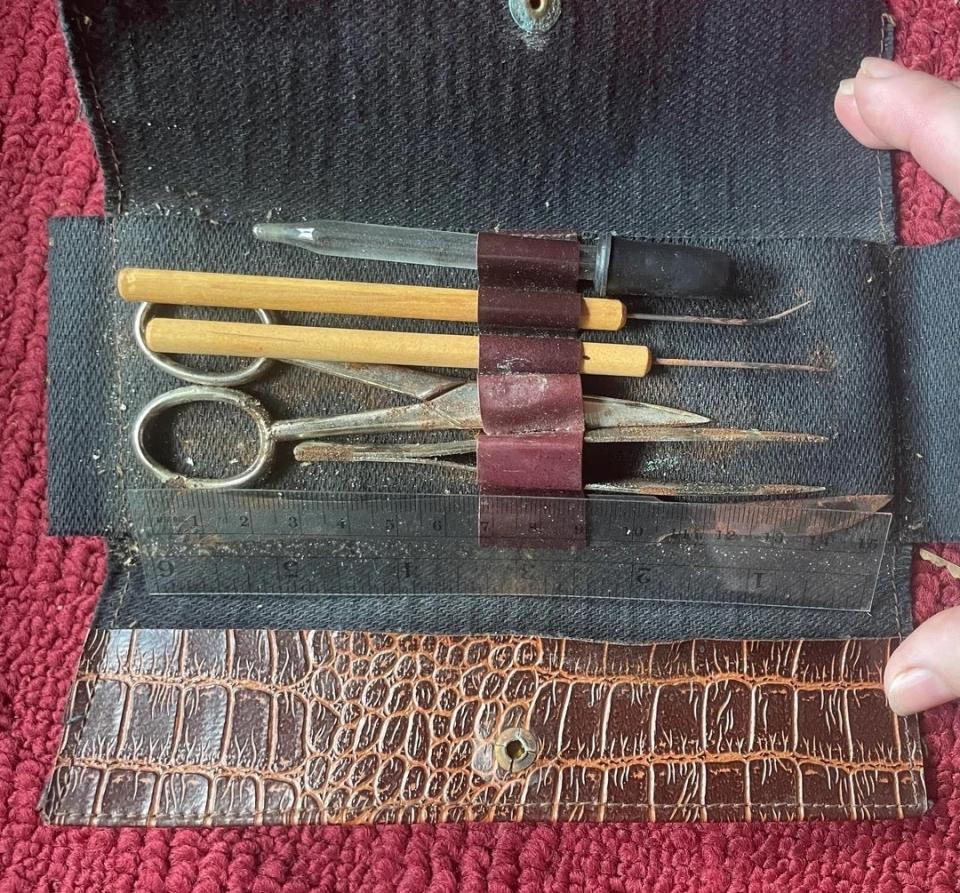 A weathered leather case holds a variety of vintage manicure tools, including a nail file, scissors and cuticle pushers, on a textured red surface