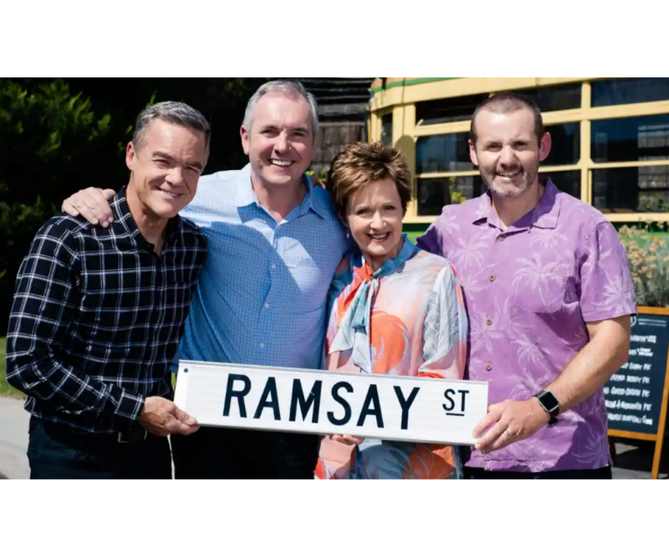 Some of the cast of Neighbours stand smiling holding the iconic Ramsay St sign. 