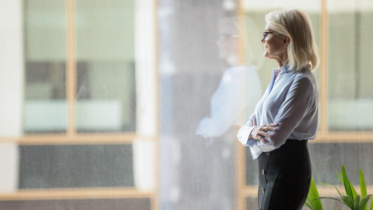 Smiling confident mature businesswoman pondering strategy, standing near big window in modern office, looking at cityscape, entrepreneur lost in thoughts, planning future project, business vision.