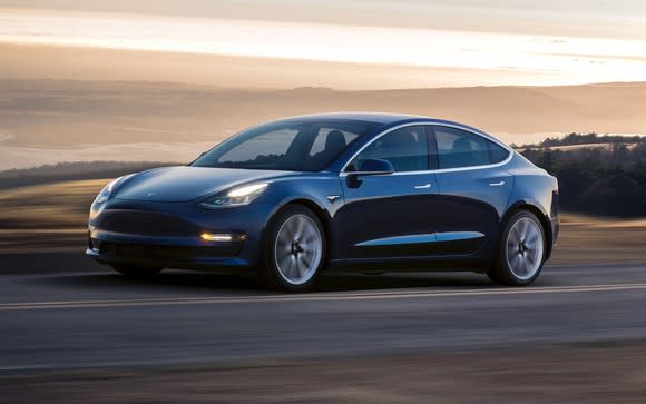 Blue Tesla Model 3 driving on an open road on a rolling prairie in front of a hazy background.
