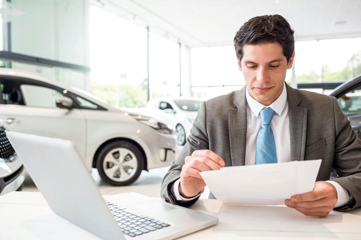 Male car dealer reading over a document while sitting at desk in front of laptop in car dealership showroom with cars for sale in background