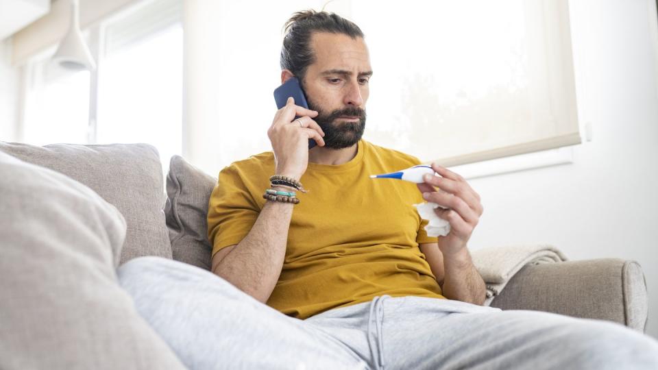 worried man talking on smart phone while looking at thermometer at home