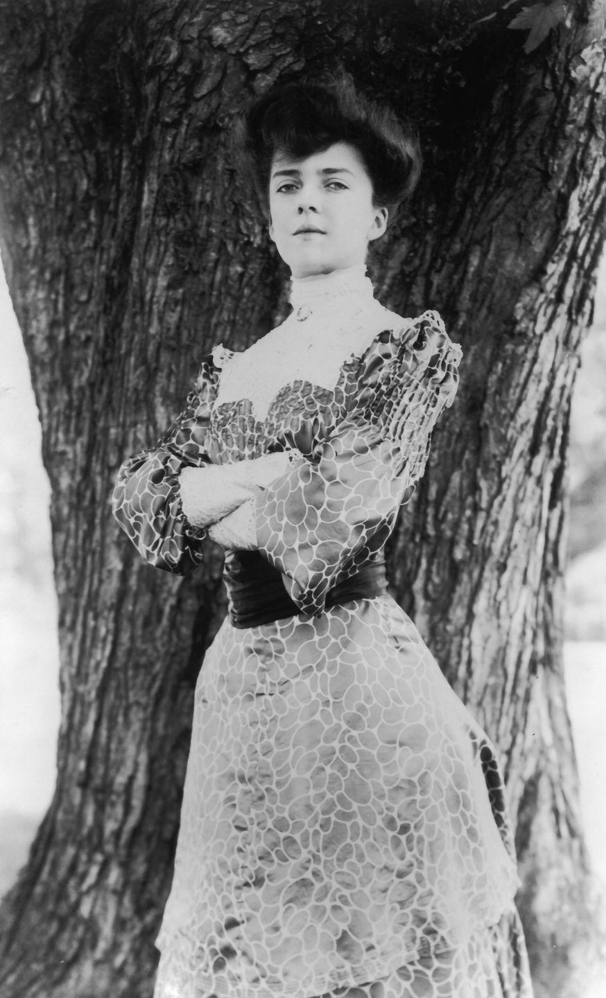 a black and white photo of alice roosevelt, wearing a dress and folding her arms while standing in front of a tree