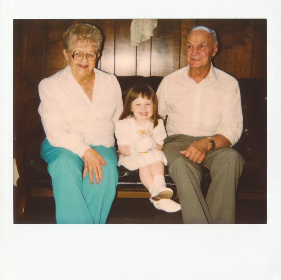 An elderly woman and man sit on a bench with a young girl between them, all smiling warmly at the camera