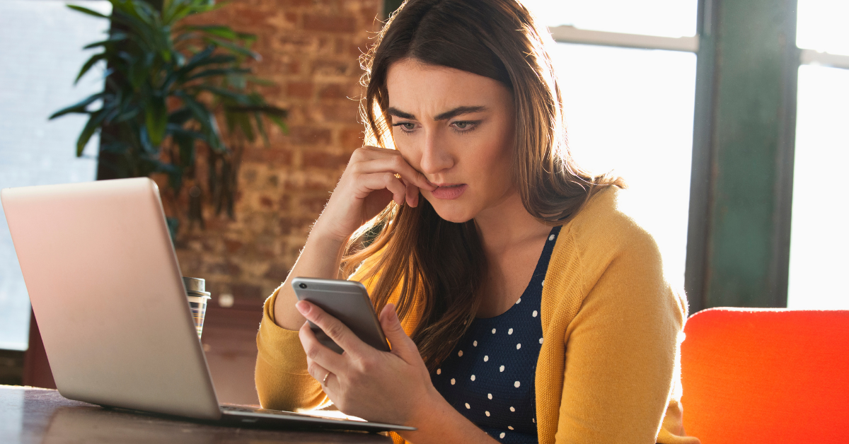 A young woman looks concerned as she holds her phone with an open laptop in front of her.