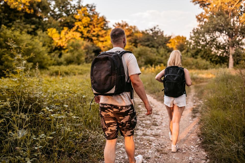 a man and a women wearing backpacks and walking on a gravel path through the woods