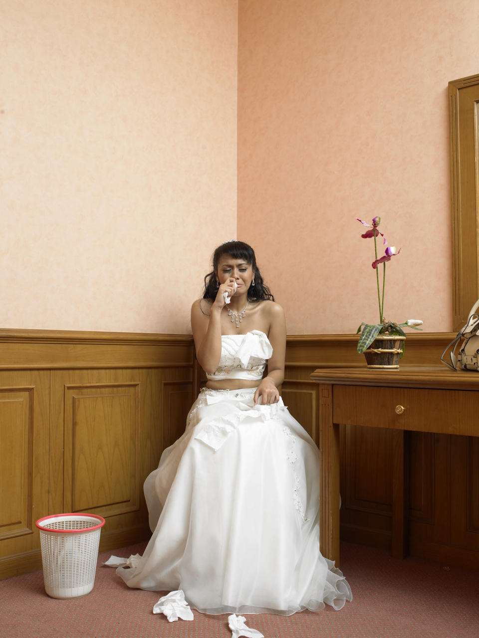 Bride sitting, emotional, in a white wedding dress, tissue in hand, indoors with simple decor