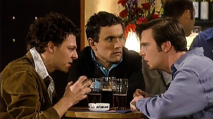 Three guys huddled around a barstool with drinks talking in a scene from Coupling.