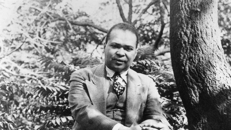 poet countee cullen sitting on a large rock next to a tree trunk and crossing his hands over a knee