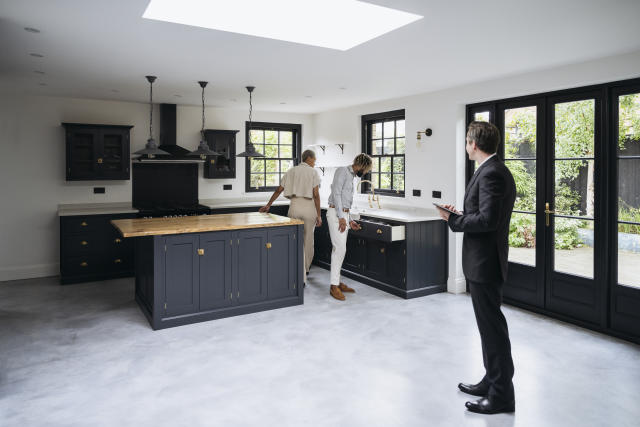 Wide angle view of real estate agent watching as Black couple in 20s and 30s check details of Shaker-style kitchen with view to outdoor patio and garden.