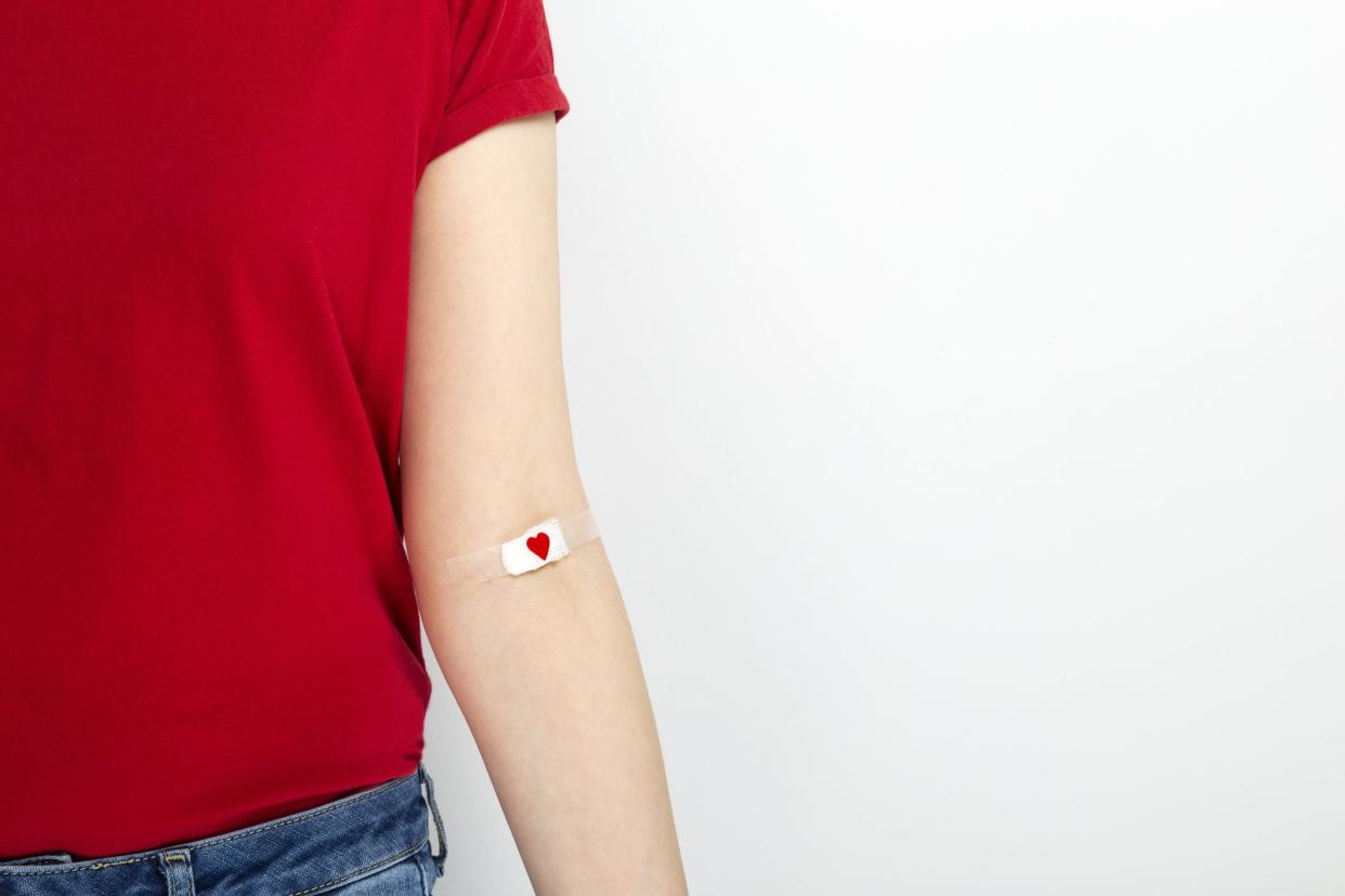 Blood donorship. Young girl in red T-shirt hand taped with patch with red heart after giving blood on gray background. Copy space