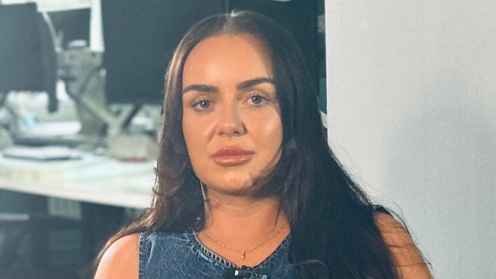 Haileigh has long brown hair. She is sitting in her office and looking straight into the camera. She is wearing a gold necklace and a denim top.