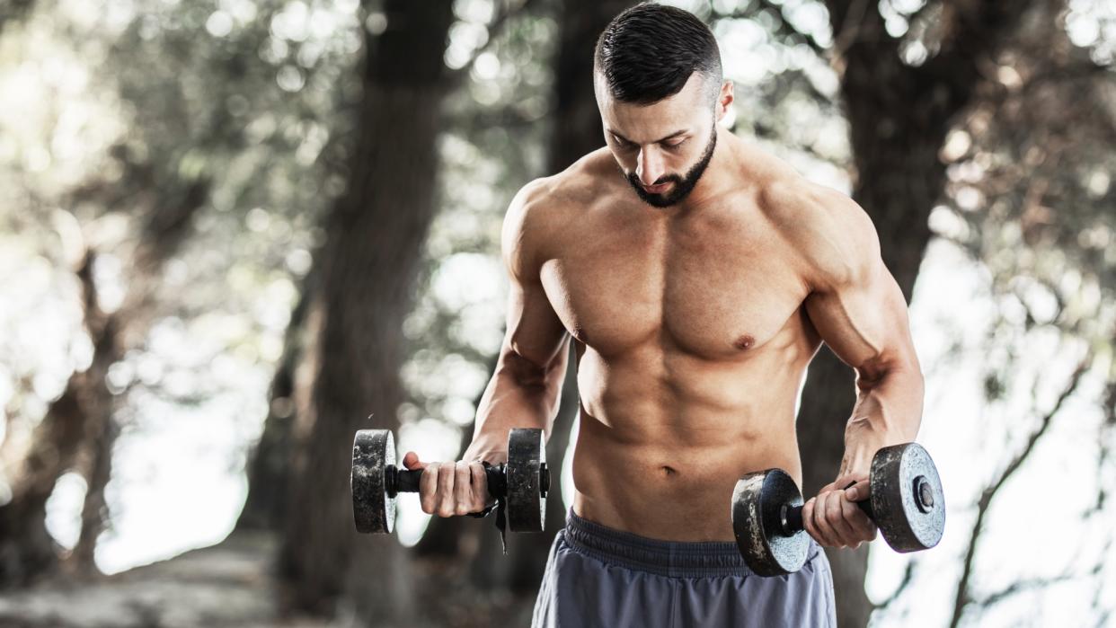  Man holding two dumbbells during workout standing outdoors in nature. 