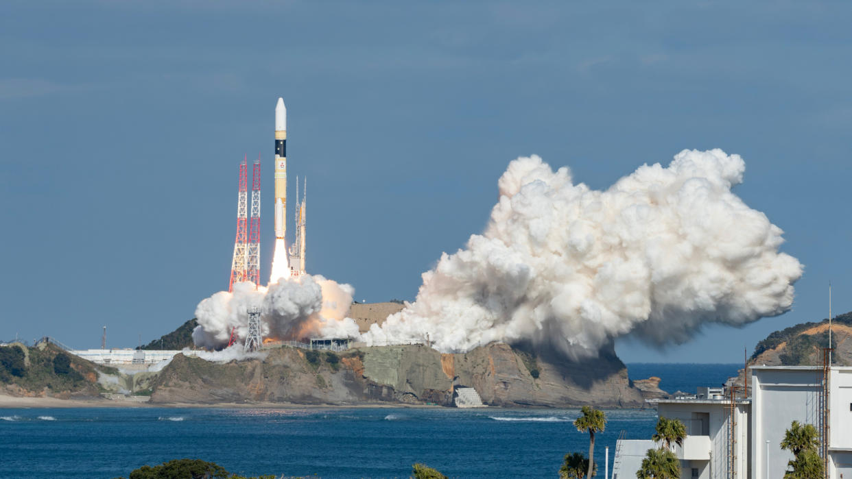  A white, black and orange rocket lifts off a seaside launch pad, creating a large plume of white exhaust. 
