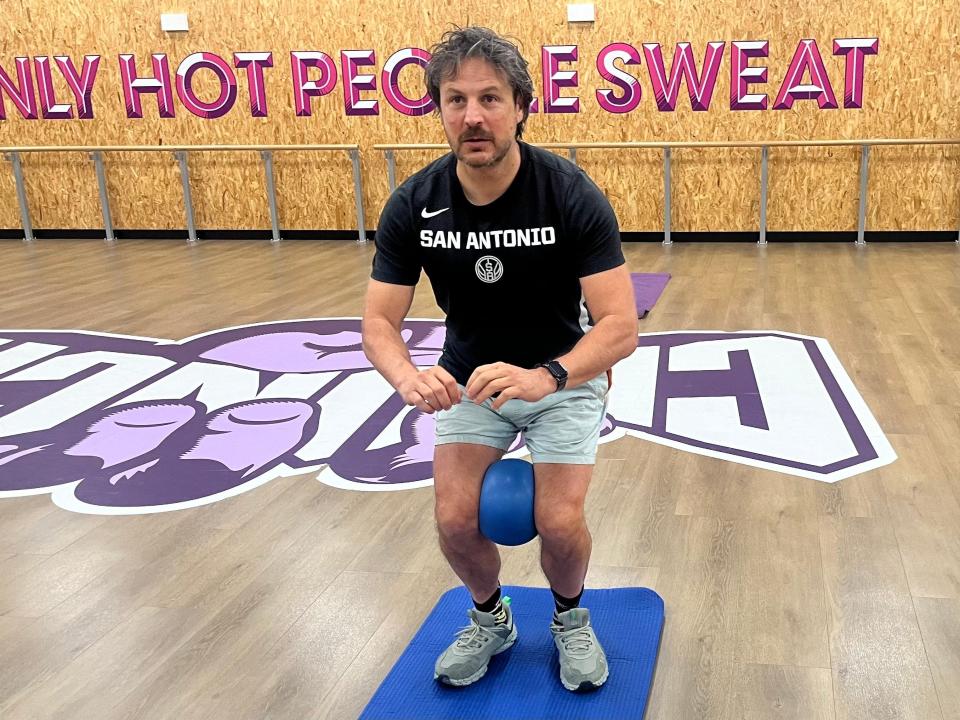 The writer stands on a blue Pilates mat and crouches with a blue ball between his knees. He stands in a gym with wooden floors and pink lettering in the background