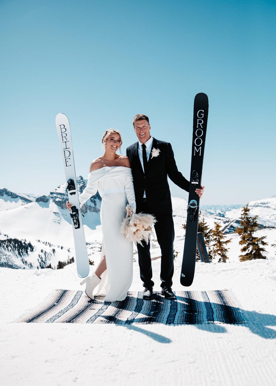 A bride and groom pose for a photo on top of a snowy mountain holding custom skis.