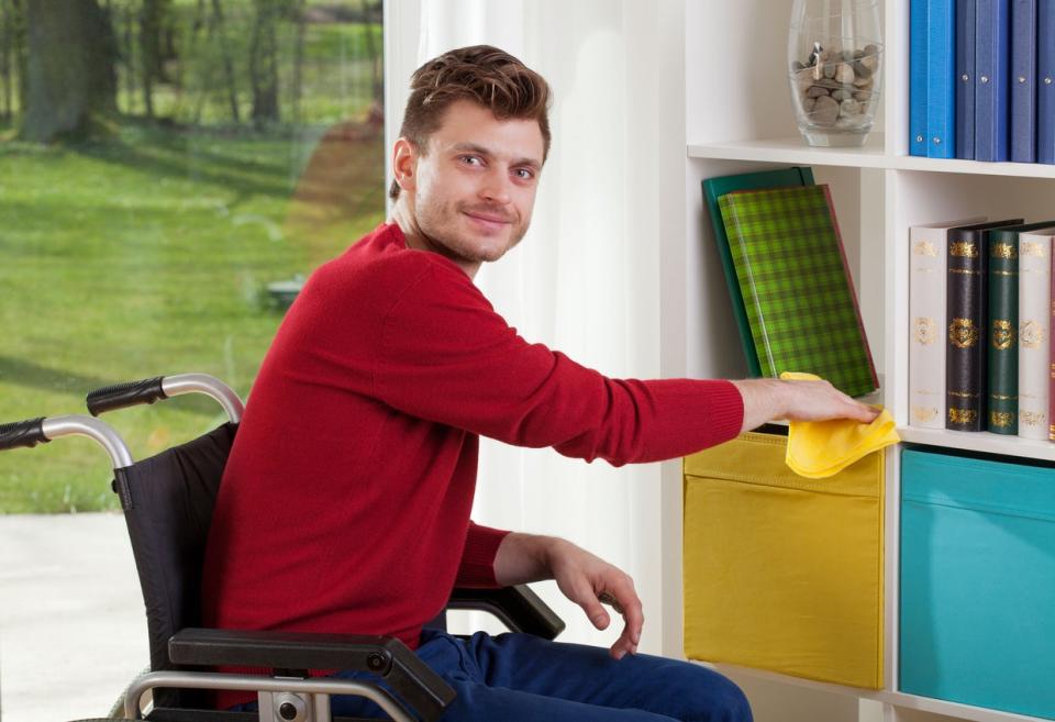 Man in wheelchair dusting shelves at home.