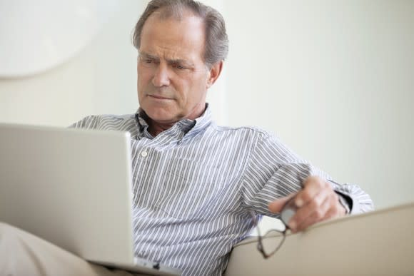 An older man reads material on his laptop while sitting on a couch and holding his glasses in his hand.