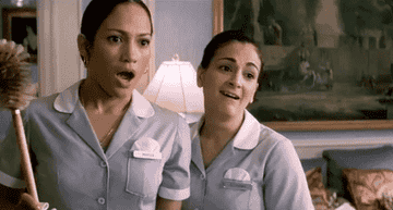 Jennifer Lopez and a co-worker in maid uniforms, surprised and smiling, in a hotel room setting. Jennifer holds a duster, and both have name tags