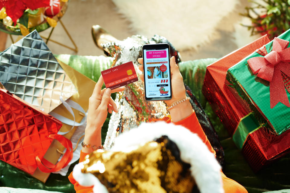 Holiday season. female in sparkle gold sequin santa hat with credit card browsing high fashion retail online store and on a smartphone while sitting on divan in the modern living room at Christmas.