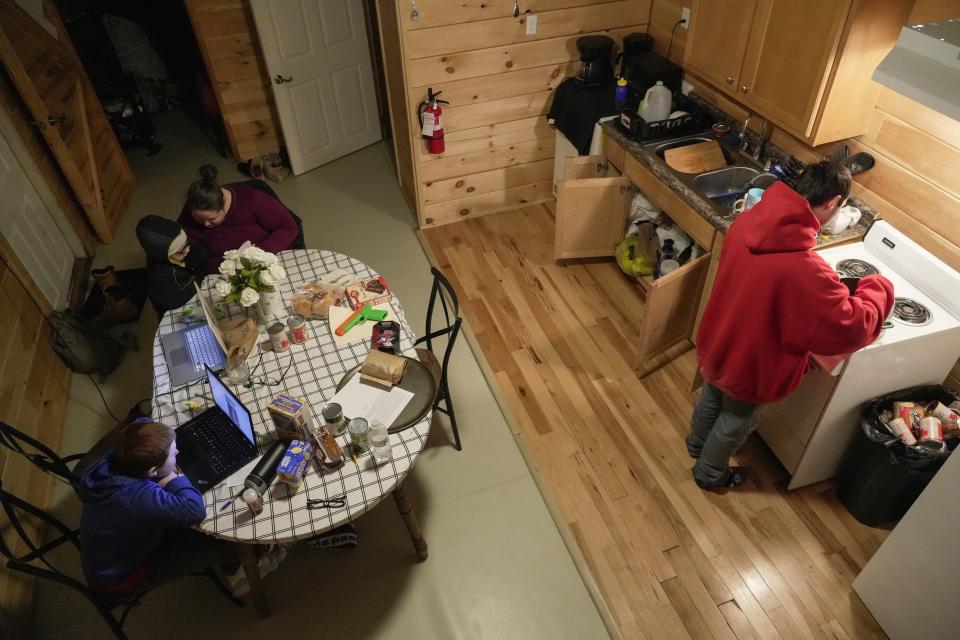 Dylan Prince, right, heats up dinner as, Ashley, top left, talks with her son Waylon while her other son, Hunter watches a video on his computer inside the family's temporary home at Camp Graves, Tuesday, Jan. 16, 2024, in Water Valley, Ky. Two years after the tornado outbreak that killed dozens and leveled much of the real estate in Mayfield, many people, like the Prince family, are still living through another, slower disaster, the search for housing. (AP Photo/Joshua A. Bickel)