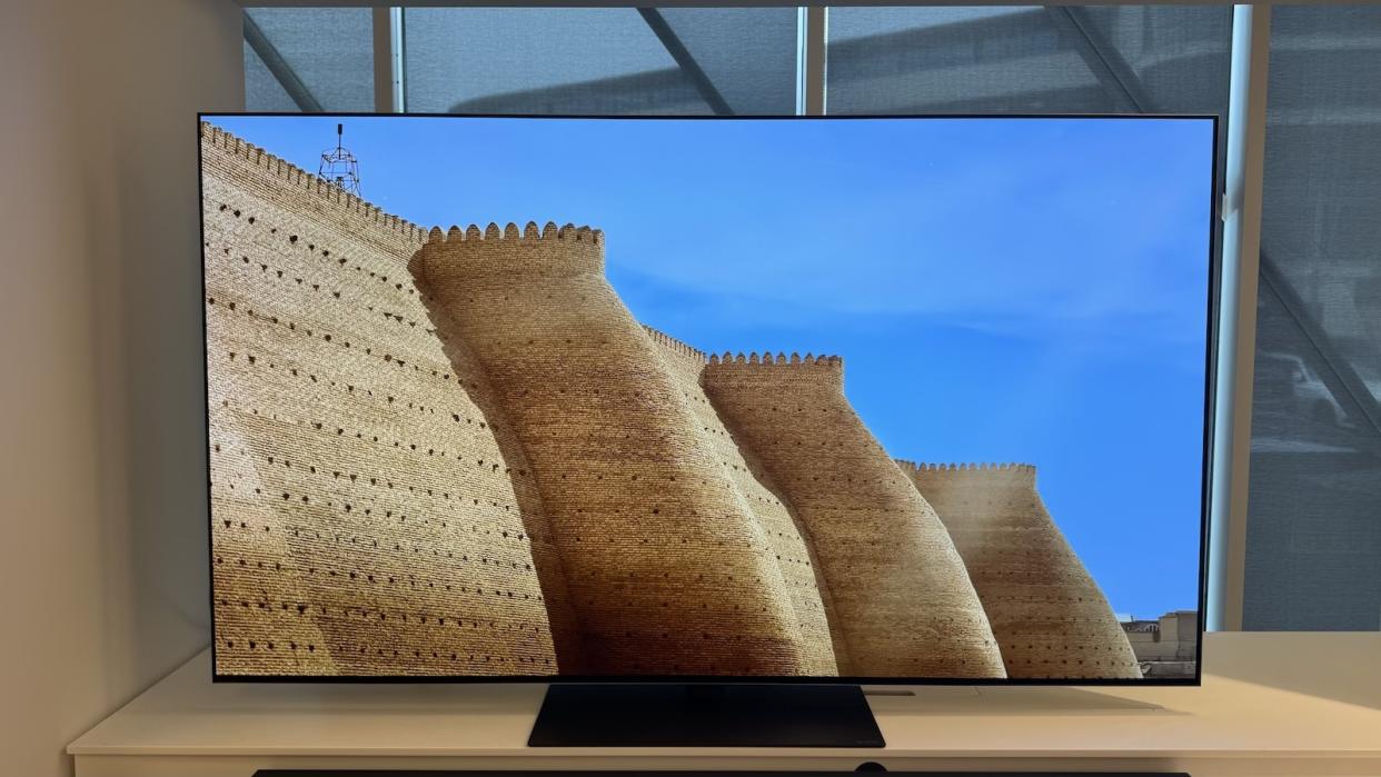  The LG G4 OLED TV photographed on a white stand in a showroom, with a soundbar positioned in front. 