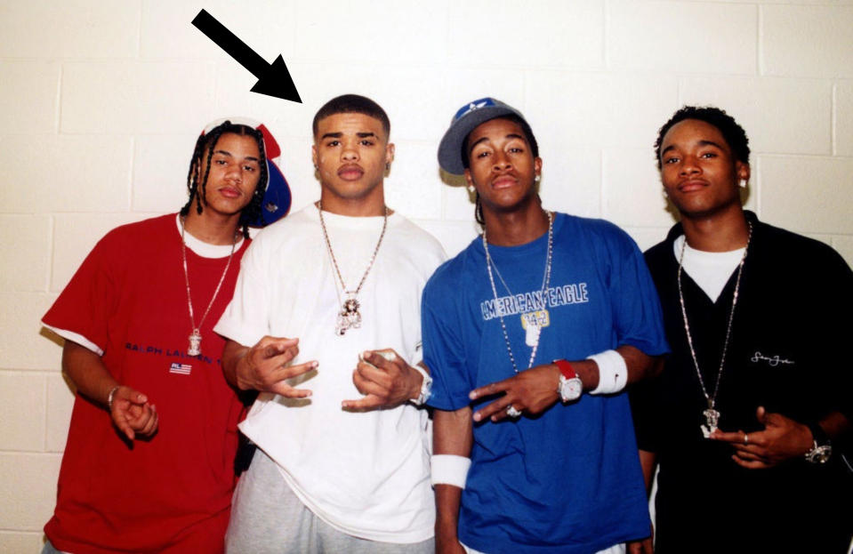B2K posing for a photo in front of a concrete wall with an arrow pointing at Raz B who is second from the left wearing a tall T and sweatpants