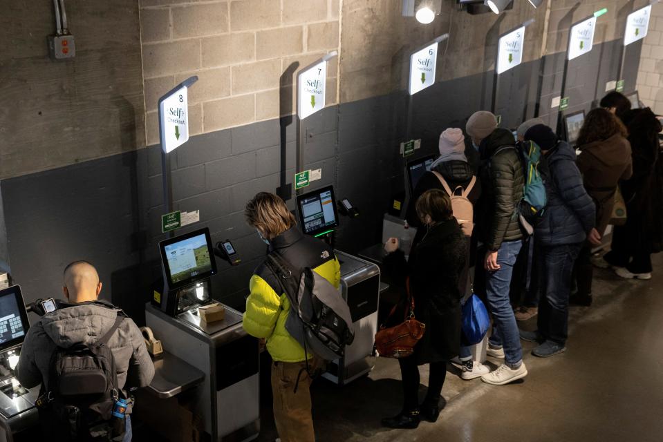 People wearing coats and other cold-weather clothing scan and pay for groceries at a supermarket in New York City. Lighted signs that read "Self-checkout" stand above each checkout kiosk.