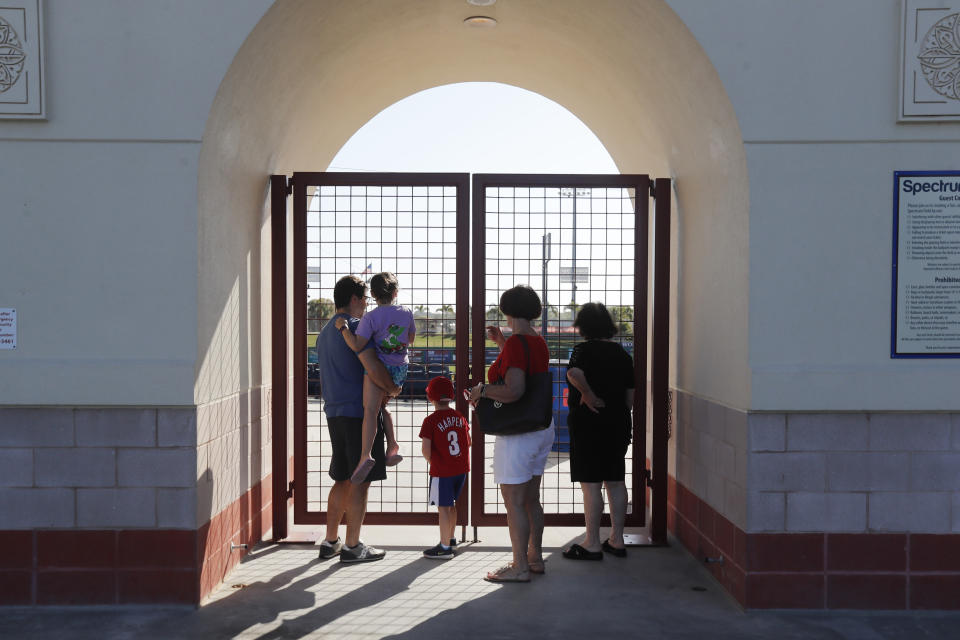 Fans look from outside the gates to Spectrum Field, spring training baseball game home of the Philadelphia Phillies which are locked, Friday, March 13, 2020, in Clearwater, Fla. League Baseball has delayed the start of its season by at least two weeks because of the coronavirus outbreak and suspended the rest of its spring training schedule. (AP Photo/Carlos Osorio)