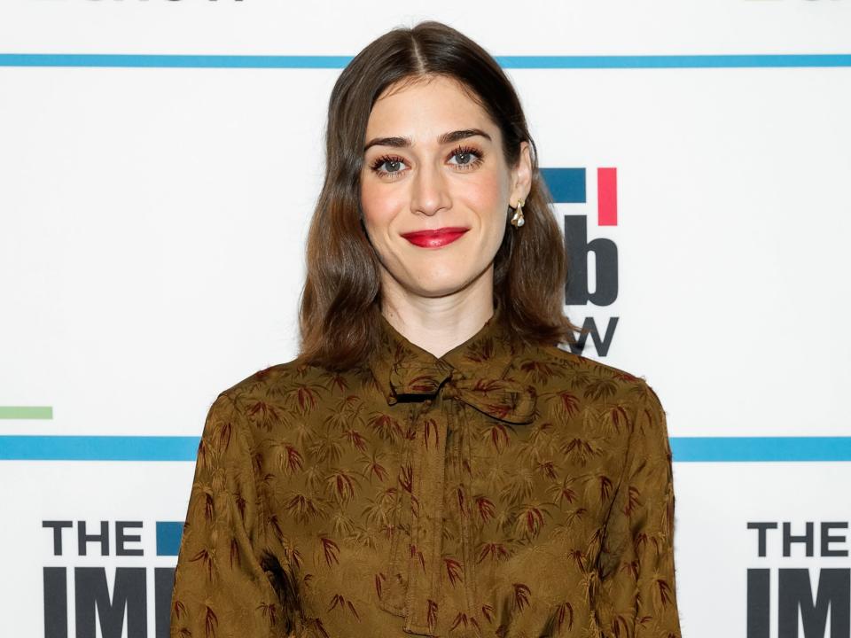 Lizzy Caplan in a brown dress with a collar wearing red lipstick and blush in front of white background