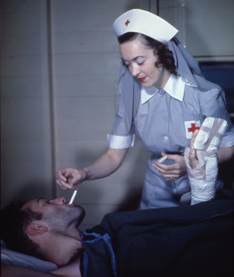 A nurse in a vintage uniform assists a male patient with a bandaged hand, offering him a cigarette while he lies in bed