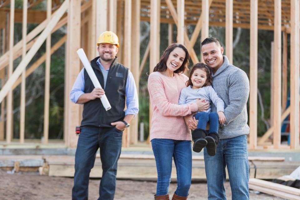 A family and construction worker smile while standing in front of their future house which is in the early stages of being built.