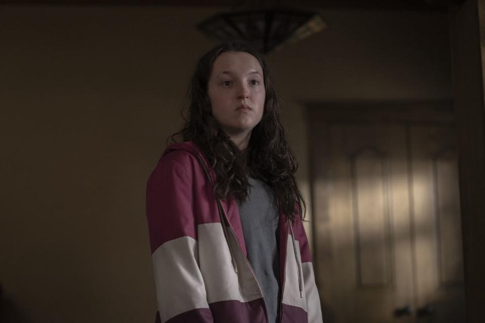 bella ramsey stands in character in a room during a season 1 episode of the last of us, she wears a red and white stripped zip up jacket and gray shirt