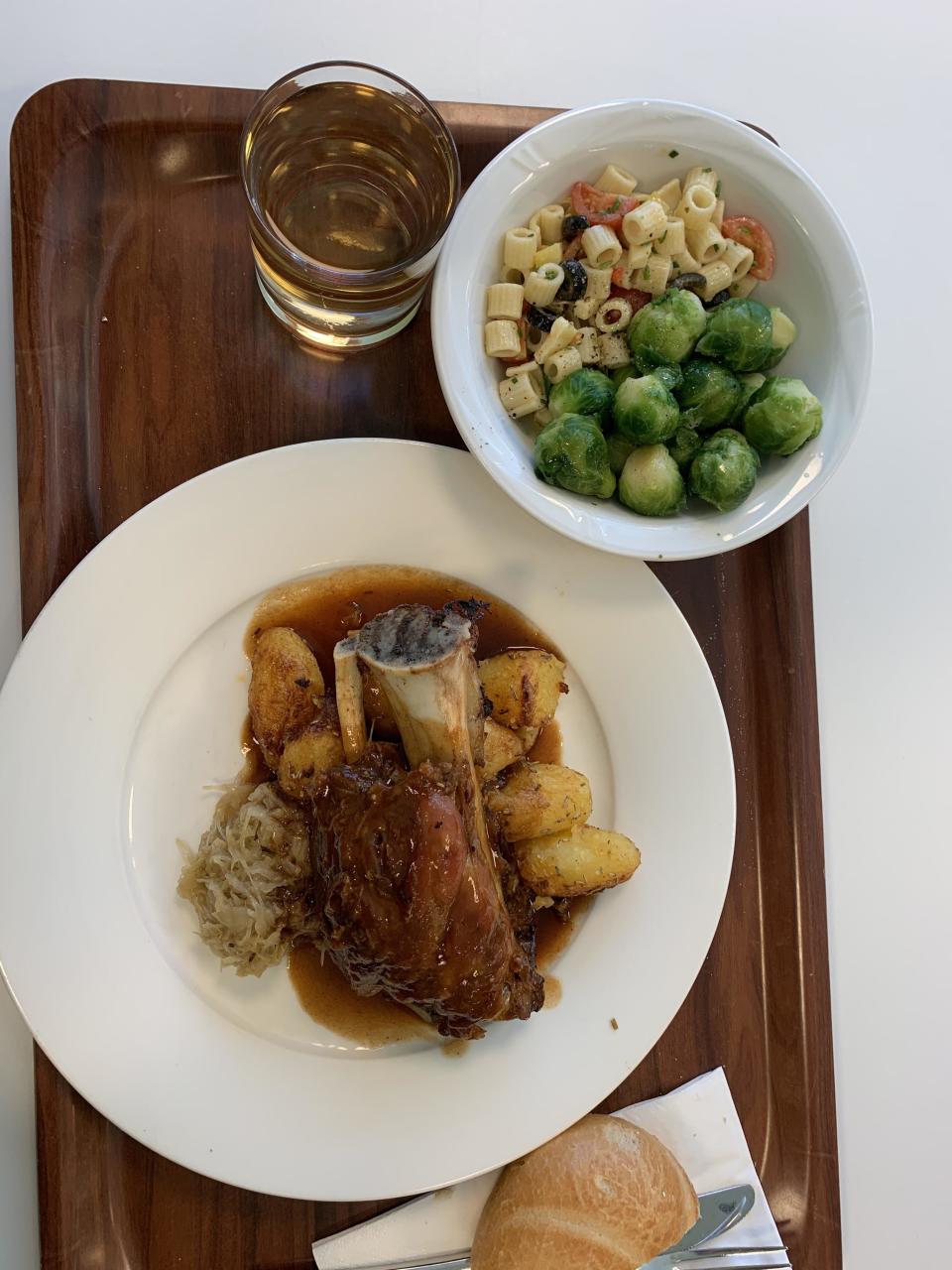 A plate with a large piece of meat and potatoes, with Brussels sprouts, pasta, and a roll on the side