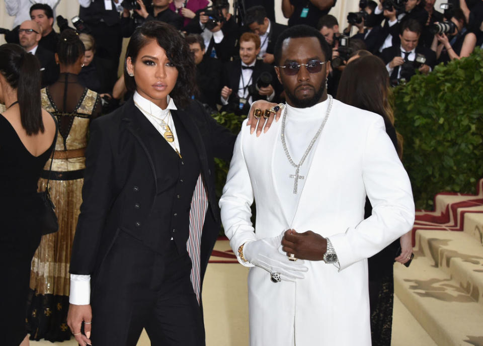 Cassie Ventura in a black suit stands next to Sean "Diddy" Combs in a white suit at a red carpet event