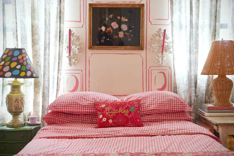 Floral art and candle sconces above bed with red bedding flanked by lamps in white bedroom with with trompe l'oeil detail.