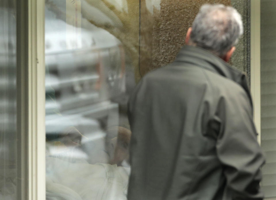 In this March 12, 2020, photo, Steve Sedlacek, right looks at his father, Chuck Sedlacek, back left, through reflections in the window of his room at Life Care Center in Kirkland, Wash., near Seattle. The facility has been at the center of the coronavirus outbreak in the state, and Sedlacek's brother Scott said that he and his siblings have barely spoken to their father inside the center, who in addition to testing positive for the coronavirus, has blindness, neuropathy, and has difficulty using a phone, saying he is more of an "inmate" than a patient. Residents of assisted living facilities and their loved ones are facing a grim situation as the coronavirus spreads across the country, placing elderly people especially at risk. (AP Photo/Ted S. Warren)