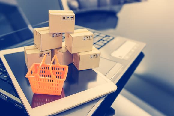 A stack of miniature boxes and a mini shopping basked on top of a tablet and laptop.