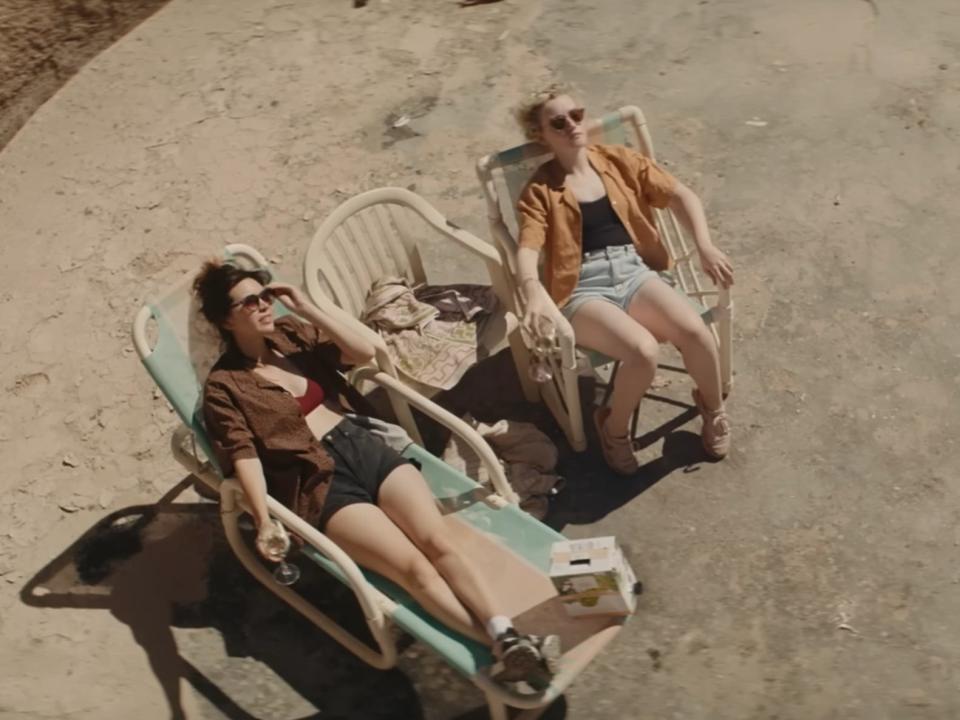 Jessica Henwick as live and Julia Garner as hanna in a scene from the royal hotel sitting on lawn chairs in the desert