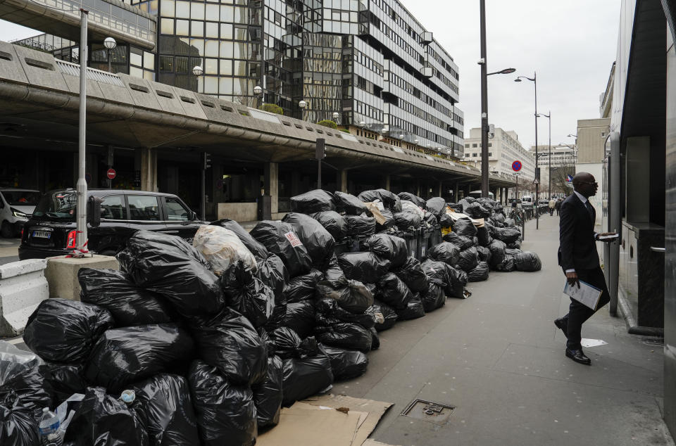 A man walks past uncollected garbages in Paris, Friday March 17, 2023, as sanitation workers are on strike. Protests against French President Emmanuel Macron's decision to force a bill raising the retirement age from 62 to 64 through parliament without a vote disrupted traffic, garbage collection and university campuses in Paris as opponents of the change maintained their resolve to get the government to back down. (AP Photo/Lewis Joly)