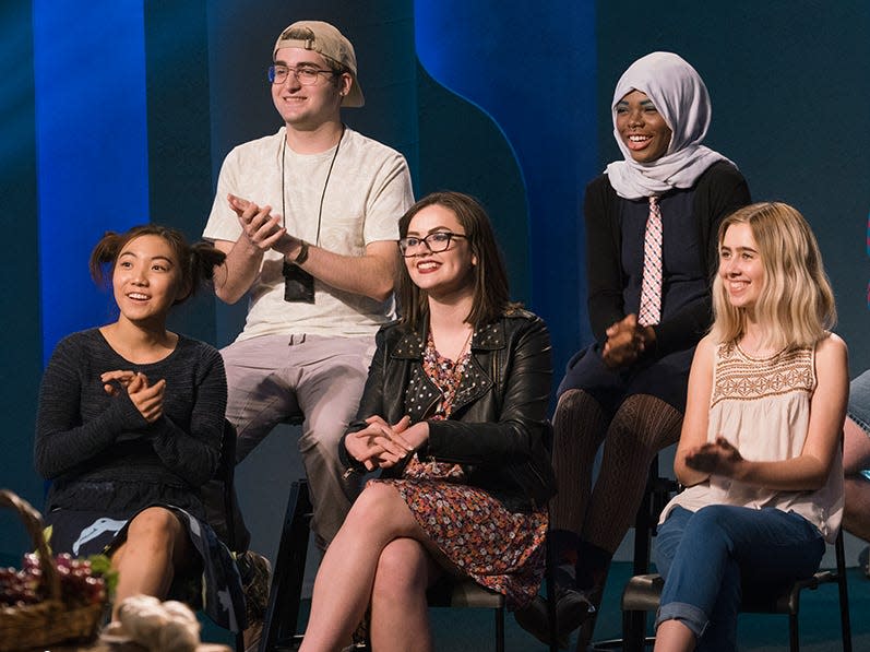 Seven contestants sit next to runway in blue room. The writer is second from the left and is smiling